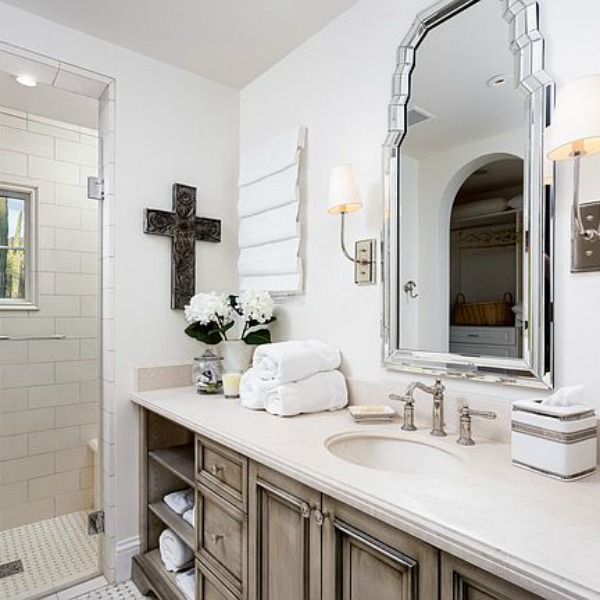 Beautifully designed French farmhouse white bathroom with antiqued vanity and cross on wall. #frenchcountry #bathroomdesign #whitebathroom