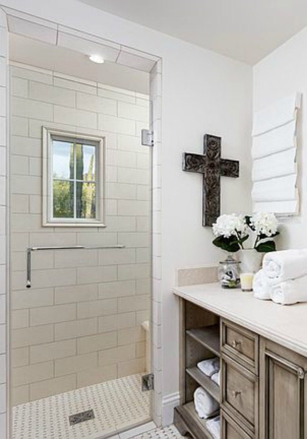 Beautifully designed French farmhouse white bathroom with antiqued vanity and cross on wall. #frenchcountry #bathroomdesign #whitebathroom