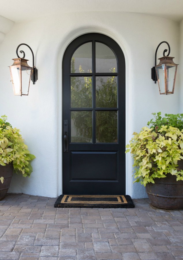 Black arched front door flanked by Old World  lanterns and white Stucco house exterior. Jaimee Rose Interiors.