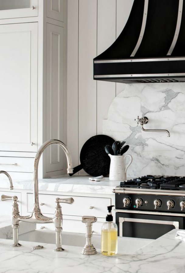 Classic white custom kitchen with black French range, marble decorative slab backsplash, farm sink, and polished nickel bridge faucet. Jaimee Rose Interiors. #kitchendesign #blackandwhite