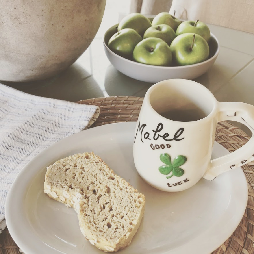 Serene tabletop with vintage Mabel mug, bowl of green apples, and homemade oat bread - Hello Lovely Studio.