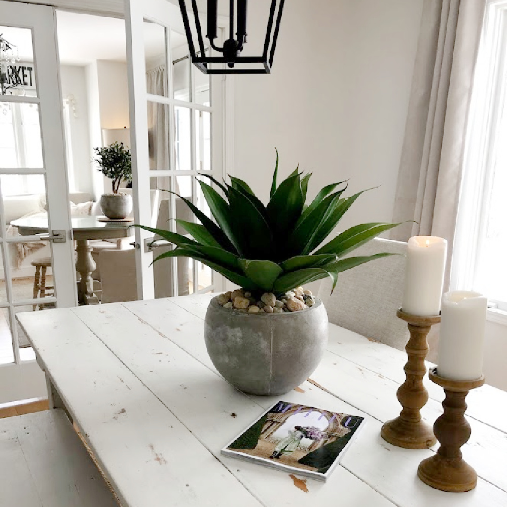 Serene white modern French dining room with farm table, rustic wood candleholders, and succulent in rustic pot - Hello Lovely Studio. #allwhitedecor #modernfrench #diningroom