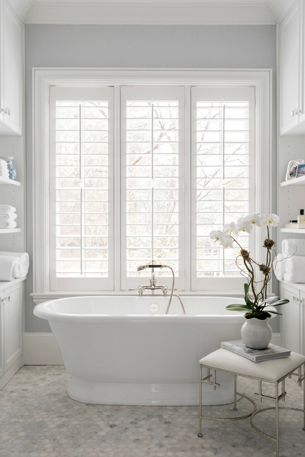 Luxurious white bathroom with freestanding tub, plantation shutters, and marble mosaic tile floor - design by Sherry Hart. #luxuriousbathroom #whitebathroom #bathroomdesign #freestandingtub #plantationshutters