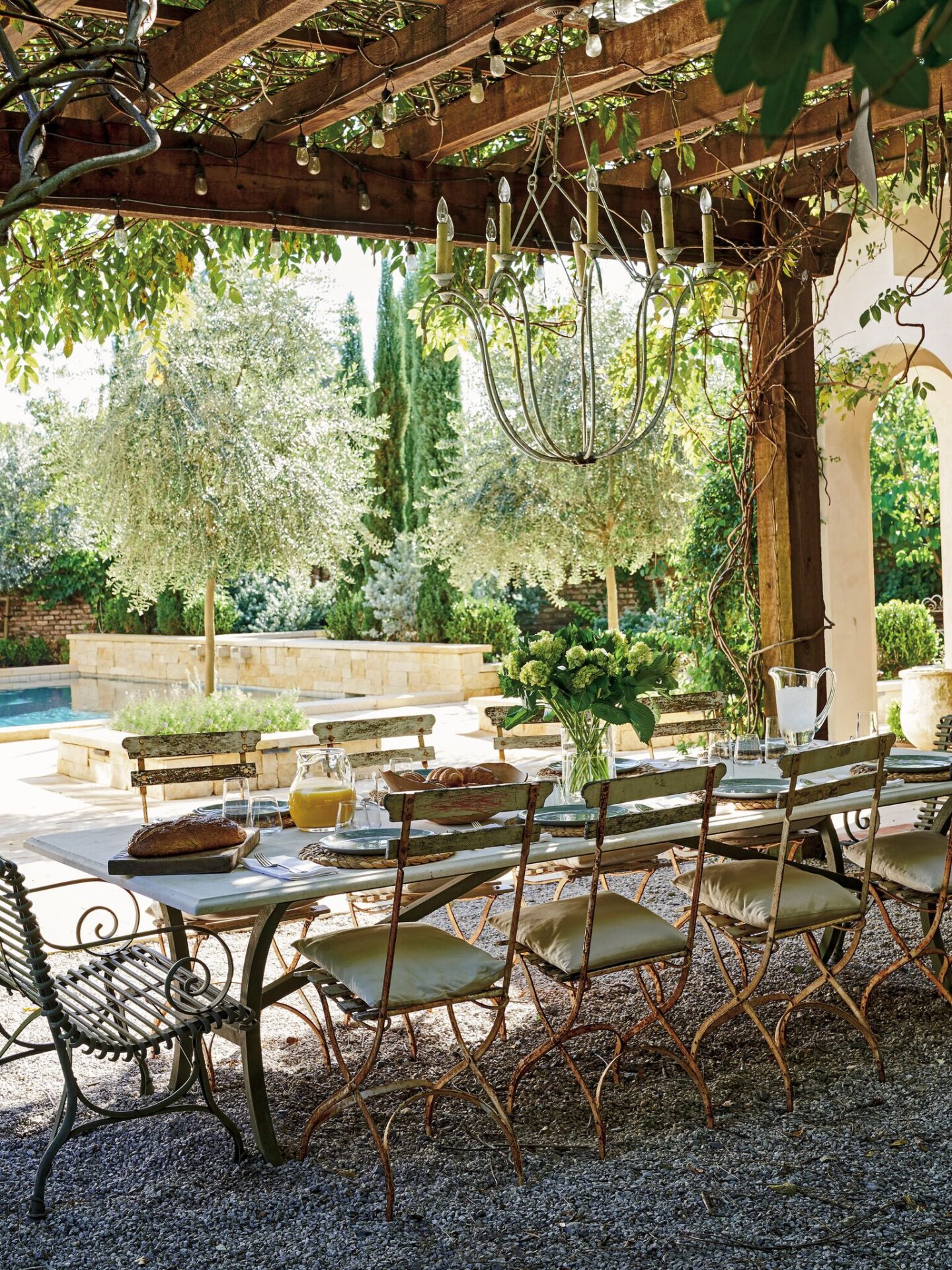 This outdoor dining area designed by Tara Shaw is an oasis with wisteria arbor, vintage tables and chairs, iron chandelier and pea gravel. #frenchgarden #frenchcourtyard #outdoordining #oldworldstyle #antiques #outdoooasis #romanticdecor