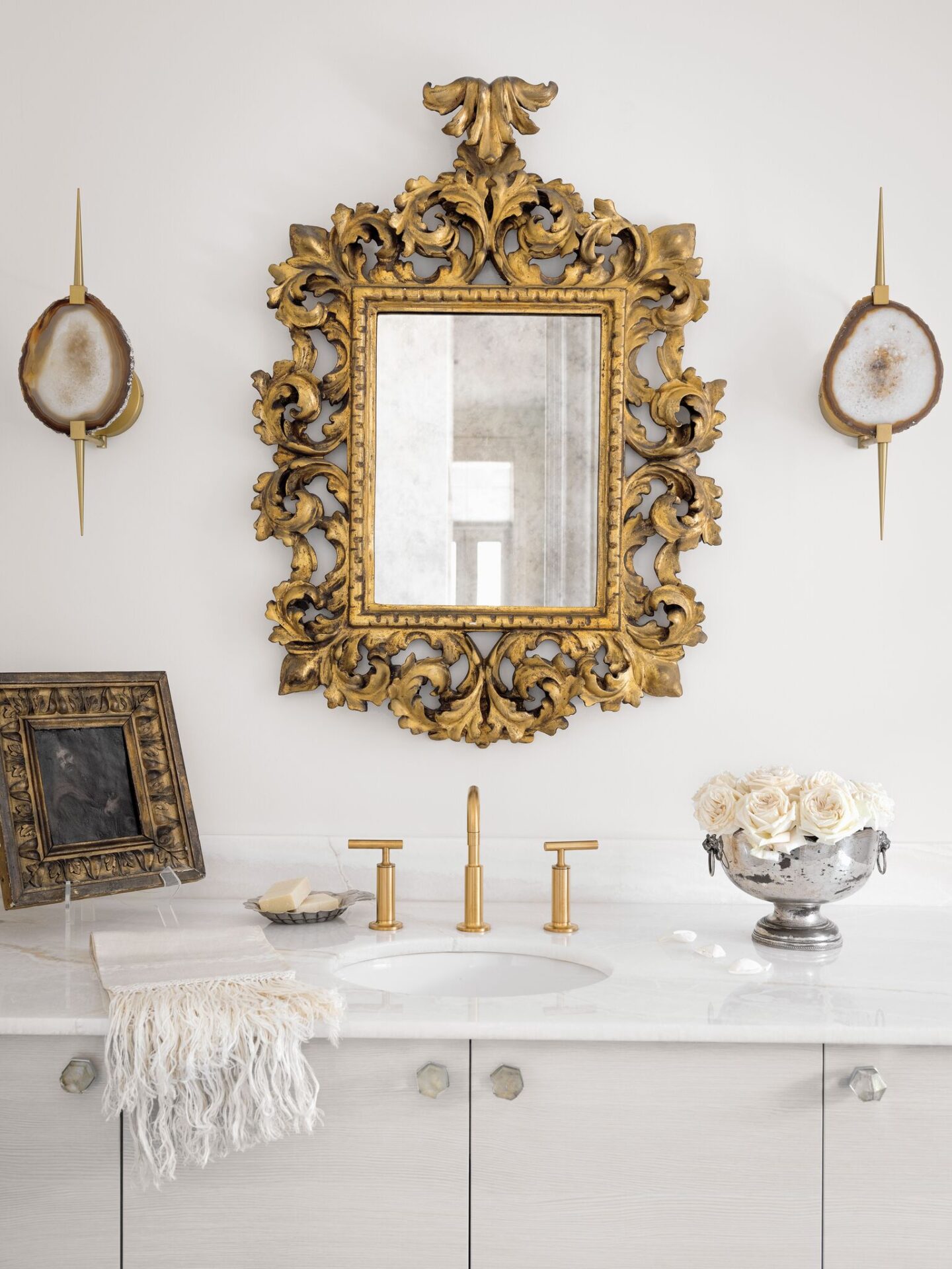 An eighteenth-century rococo Venetian mirror, contemporary onyx sconces, and gold toned fixtures harmonize in a stunningly elegant and luxurious bathroom design. #tarashaw #bathroom #interiordesign #antiques #oldworldstyle #whitebathroom #luxuriousbathroom