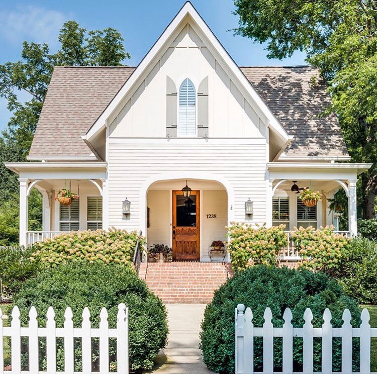 Welcoming cottage with arched entrance and front porch - Homes by Garden Gate. Charming inspiration if you love white painted house exteriors! #whitehouses #housedesign #exteriors #whitecottage #picketfence