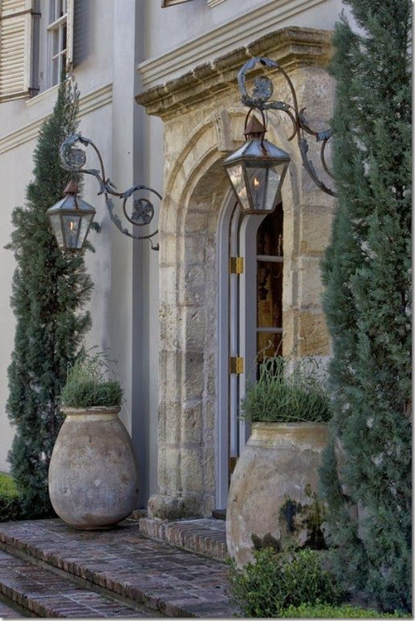 Pamela Pierce designed moment of French elegance with antique olive pots flanking an arched stone entrance with lanterns. #pamelapierce #frenchcountry #olivejars #stonesurround #frontdoor #archeddoor