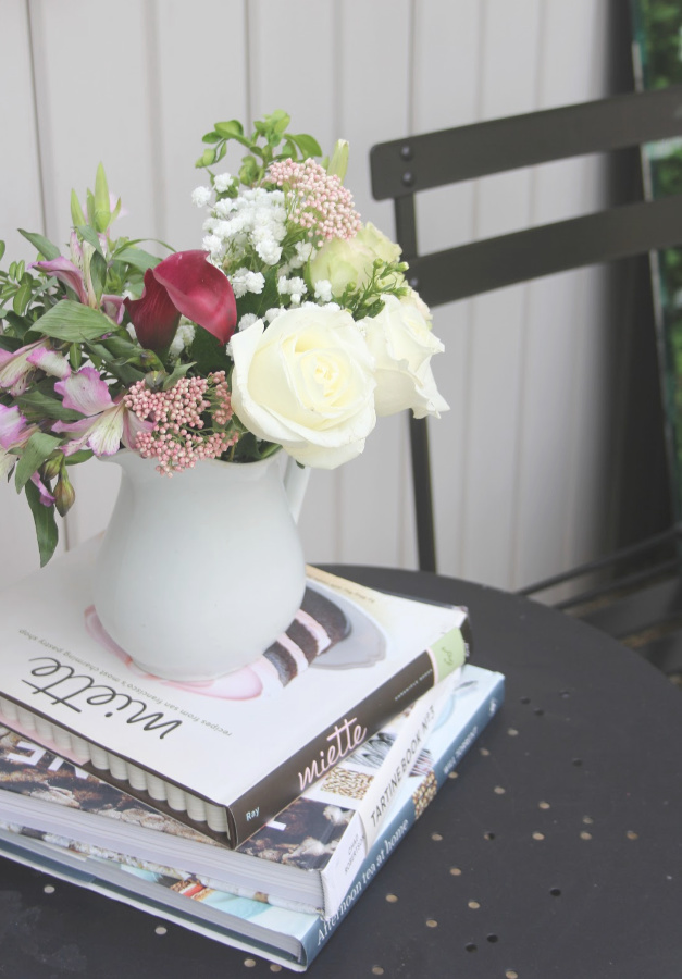 White ironstone pitcher with white roses and spring blooms atop books on a bistro table. #hellolovelystudio #ironstonepitcher #frenchcountry #florals #romanticdecor