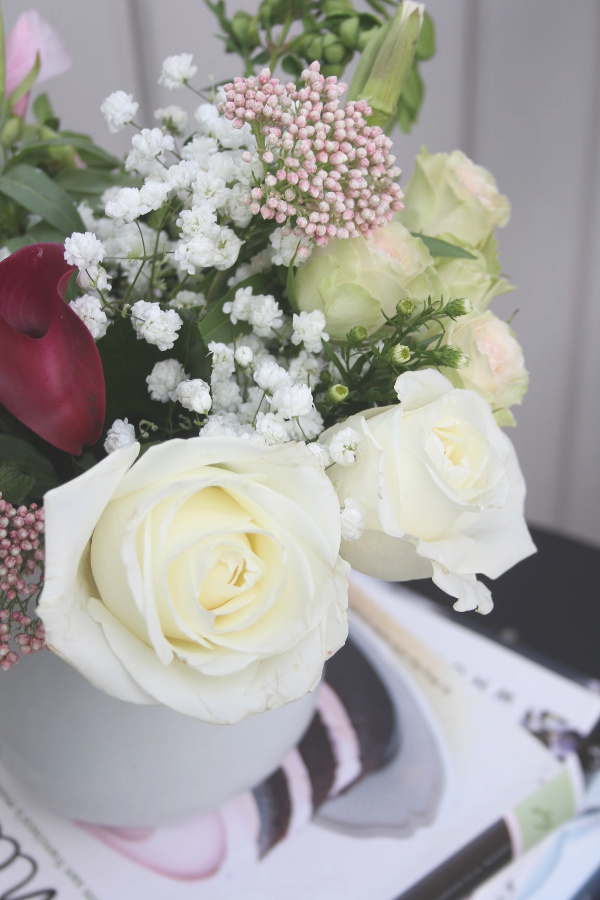 White ironstone pitcher with white roses and spring blooms atop books on a bistro table. #hellolovelystudio #ironstonepitcher #frenchcountry #florals #romanticdecor