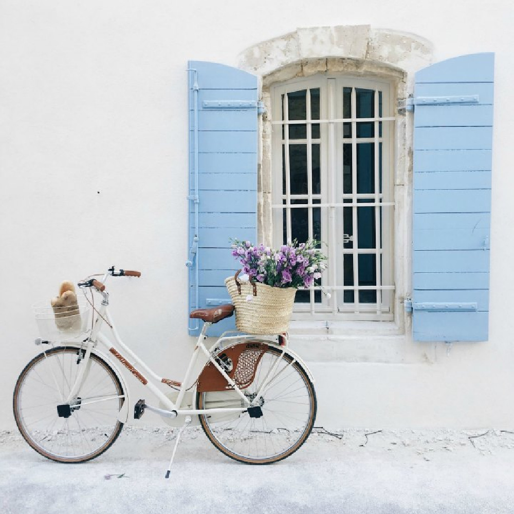 Charming French country vignette with white bicycle, French market basket of flowers, bright blue shutters, and a crisp white house exterior. Romantic indeed! Come be inspired by more French farmhouse design inspiration from Vivi et Margot on Hello Lovely. #vivietmargot #frenchfarmhouse #bicycle #frenchbasket #shutters #marketbasket #romanticdecor #frenchcountry