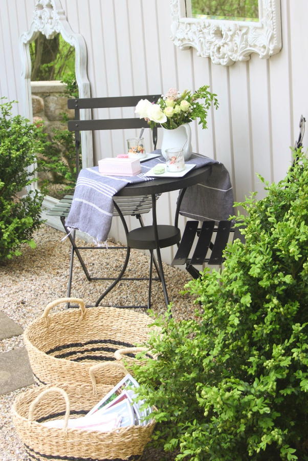 French country courtyard with boxwood, gravel, black bistro set, baskets, and a French mirror. #hellolovelystudio #cafechairs #frenchcountry #bistrochairs #outdoordining #frenchbaskets #patiofurniture #outdooroasis