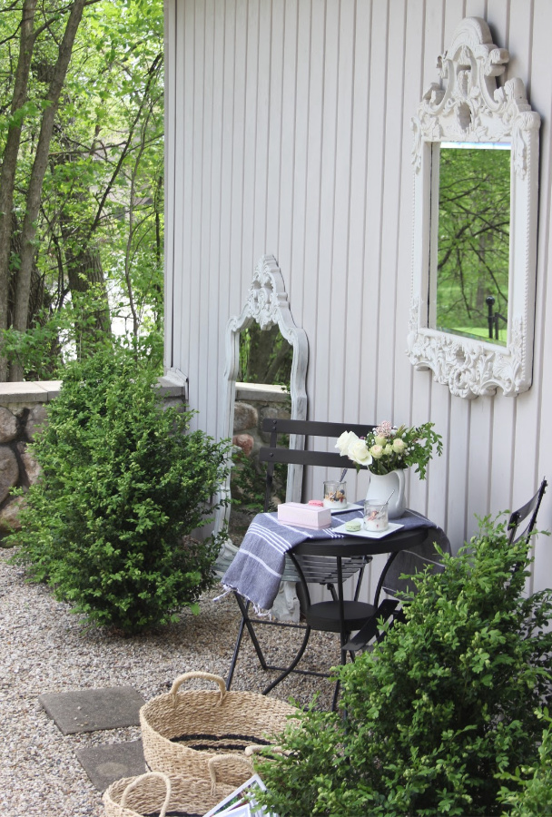 French country courtyard with boxwood, gravel, bisto set, baskets, and a French mirror. #hellolovelystudio #frenchcountry #bistroset #courtyard #gardendecor #boxwood #frenchbaskets #outdoordecor #outdooroasis