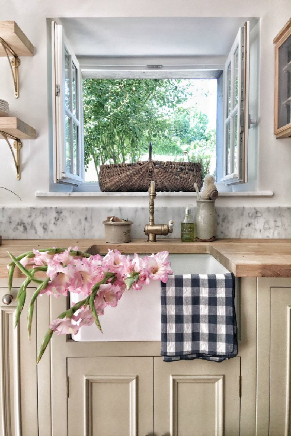 Charming French farmhouse kitchen with farm sink, checkered linens, and French baskets. A wood countertop tops putty colored cabinets. #vivietmargot #frenchkitchen #farmhousekitchen #farmsink #rusticdecor