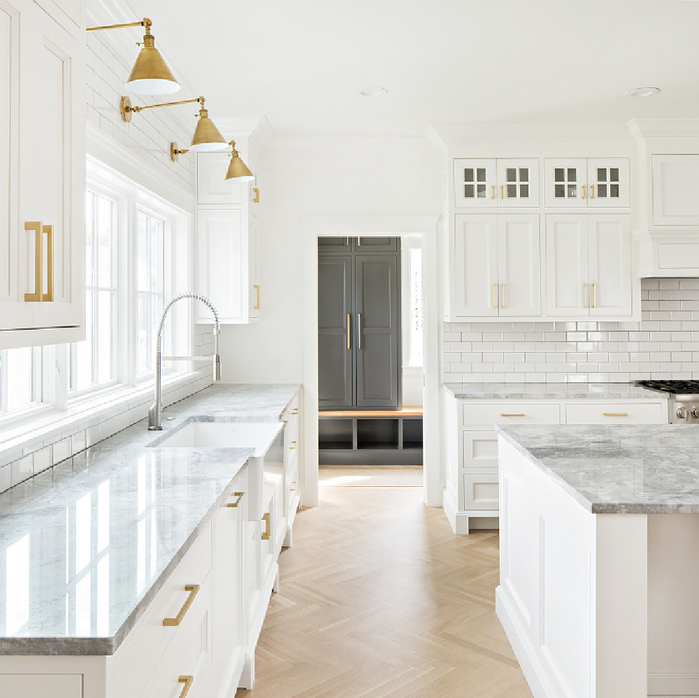 Magnificent timeless design in a white kitchen by The Fox Group. Brass library lights, white oak flooring in herringbone pattern, and quartzite countertops. Come be inspired by more Timeless Interior Design Ideas. #thefoxgroup #kitchendesign #modernfarmhouse #herringbone #classicdesign #timeless