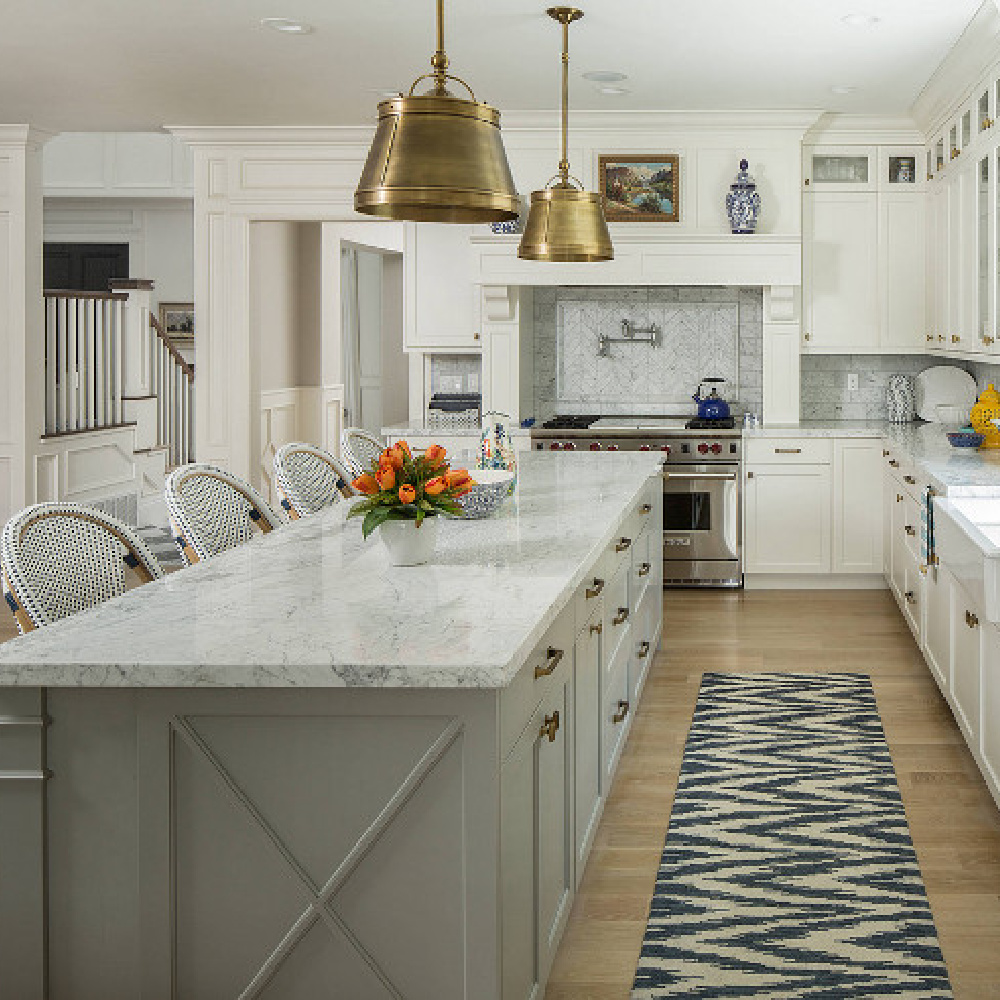 Classic kitchen by The Fox Group with brass pendant drum pendants over island and blue accents. Farm sink, white cabinetry, and Parisian counter stools. #kitchendesign #thefoxgroup #classicdesign #coastalkitchen #brasspendants