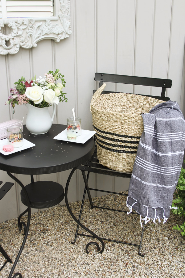 Hello Lovely French country bistro with black dining set, seagrass baskets with stripes, and Turkish stripe throw in a courtyard with gravel. #hellolovelystudio #frenchcountry #outdoordecor #bistroset #outdoordining #outdooroasis #seagrassbaskets #turkishtowel