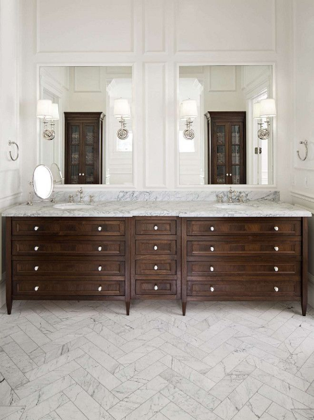 Luxurious bathroom design with marble tile in herringbone pattern. Dark wood double vanities and twin tall glass doored cabinets. Design by The Fox Group. #thefoxgroup #luxurybath #herringbonetile