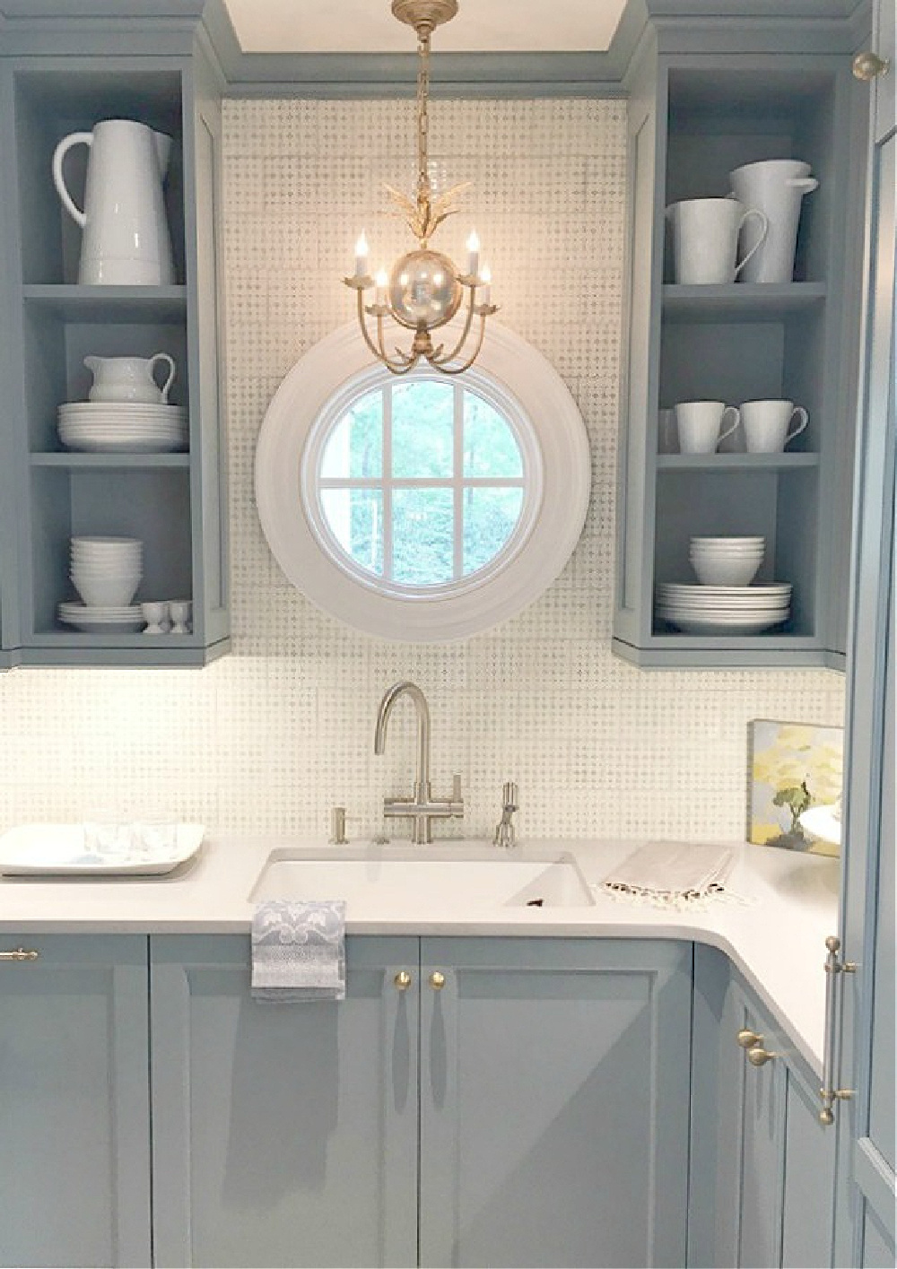 Beautiful blue butler pantry in Atlanta Southeastern Designer Showhouse 2017 kitchen . Round window and elegant brass pendant. Open shelving holds white dishware. #bluekitchen #farrowandballlightblue #timelesskitchen #kitchendesign #traditionalkitchen