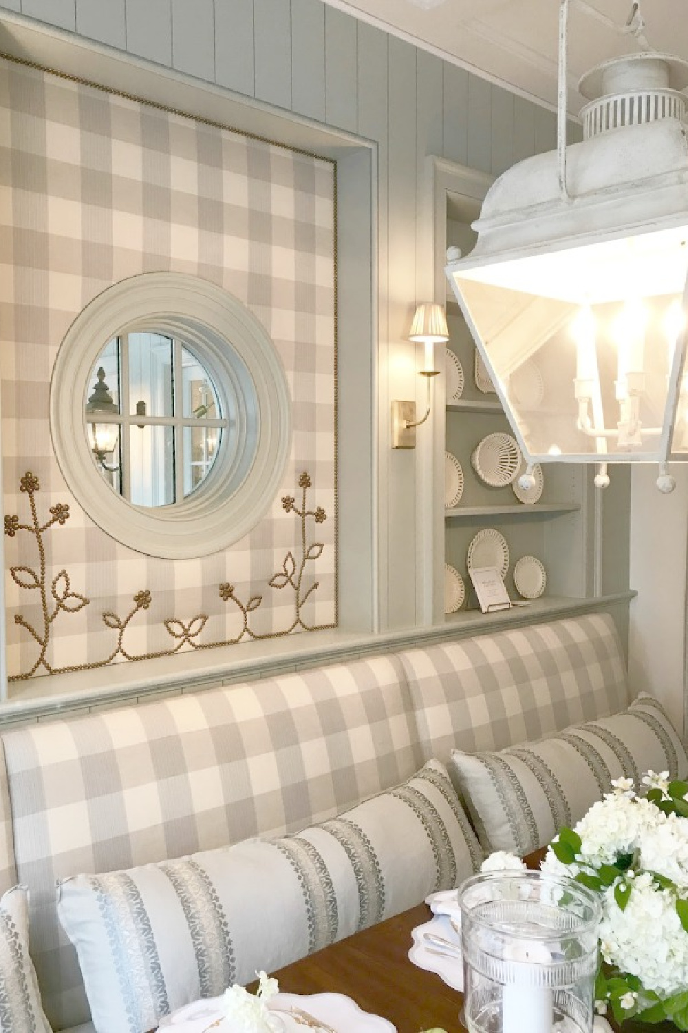 Light blue grey and cream check fabric upholstered walls and banquette in an elegant showhouse breakfast room with vintage lantern pendant. #diningroom #traditionalstyle #gingham #buffalocheck #built-ins #creamware #roundwindow #nailheadtrim #lantern #interiordesign