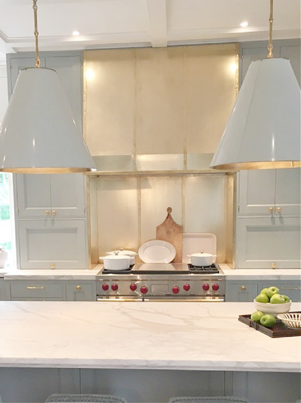 Brass custom range hood, Wolf range, light blue cabinets, and pendants over island (Circa) in a designer showhouse kitchen by Matthew Quinn. #bluekitchen #farrowandballlightblue #timelesskitchen #kitchendesign #traditionalkitchen