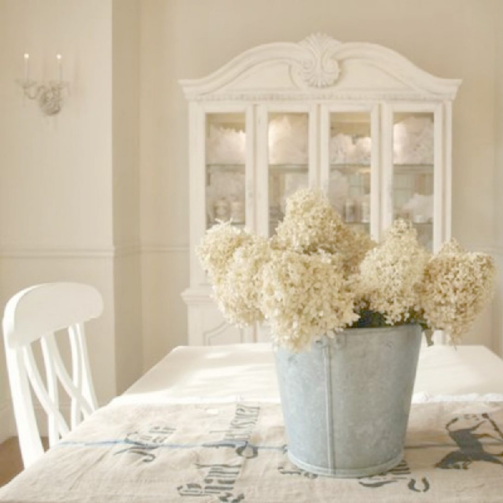 Benjamin Moore White Sand paint color on walls of my French country dining room with dried hydrangea in vintage bucket on table with grainsack runner - Hello Lovely Studio. #frenchcountry #diningroom #paintcolors #benjaminmoorewhitesand #whitesand #interiordesign