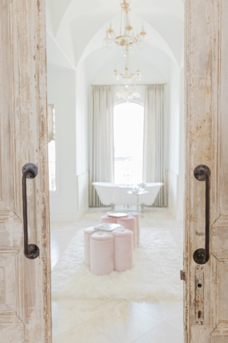 Rustic double barn doors open to an elegant French country bathroom with clawfoot tub, soaring ceilings, and a trio of chandeliers - Brit Jones. #frenchcountry #interiordesign #bathroomdesign #romanticdecor #elegantbath #luxuriousbath #barndoors #clawfoottub