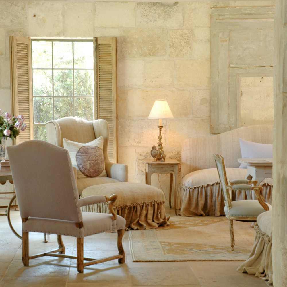 Sunny limestone, chalky hues, linen upholstered furniture with ruffled skirts, and reclaimed stone mix charmingly in a living room with French flair - Chateau Domingue. #countryfrench #interiordesign #livingroom #oldworld