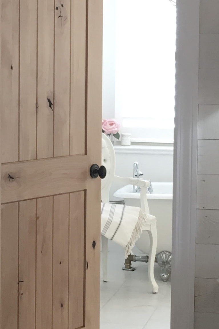 Knotty alder door to French country white bathroom by Hello Lovely Studio. #bathroomdecor #frenchcountry #whitebathroom #hellolovelystudio #alder #clawfoottub