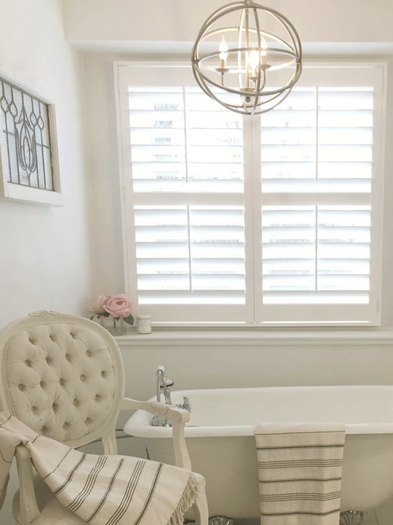 Clawfoot tub, French chair, and stripe Turkish towels in white French country bathroom by Hello Lovely Studio. #bathroomdesign #frenchcountry #hellolovelystudio #clawfoottub #plantationshutters #whitebathroom #frenchbathroom #vintagestyle