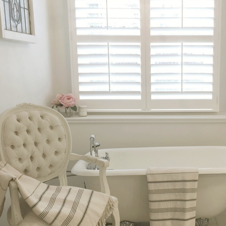 Clawfoot tub, French chair, and stripe Turkish towels in white French country bathroom by Hello Lovely Studio. #bathroomdesign #frenchcountry #hellolovelystudio #clawfoottub #plantationshutters #whitebathroom #frenchbathroom #vintagestyle