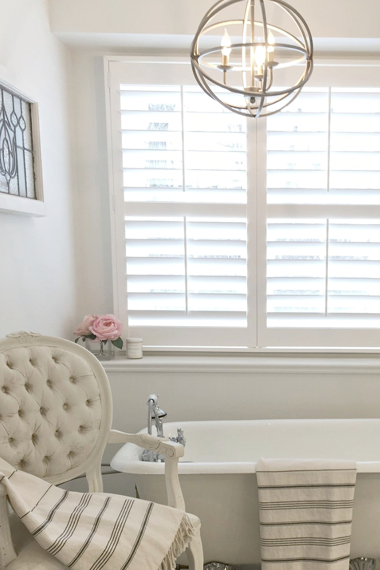 Clawfoot tub, French chair, and stripe Turkish towels in white French country bathroom by Hello Lovely Studio. #bathroomdesign #frenchcountry #hellolovelystudio #clawfoottub #plantationshutters #whitebathroom #frenchbathroom #vintagestyle