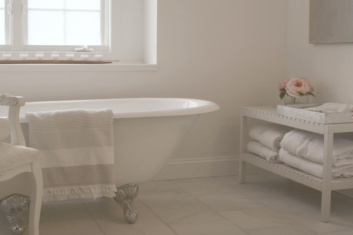 Clawfoot tub, French chair, and stripe Turkish towels in white French country bathroom by Hello Lovely Studio. #bathroomdesign #frenchcountry #hellolovelystudio #clawfoottub #plantationshutters #whitebathroom #frenchbathroom #vintagestyle