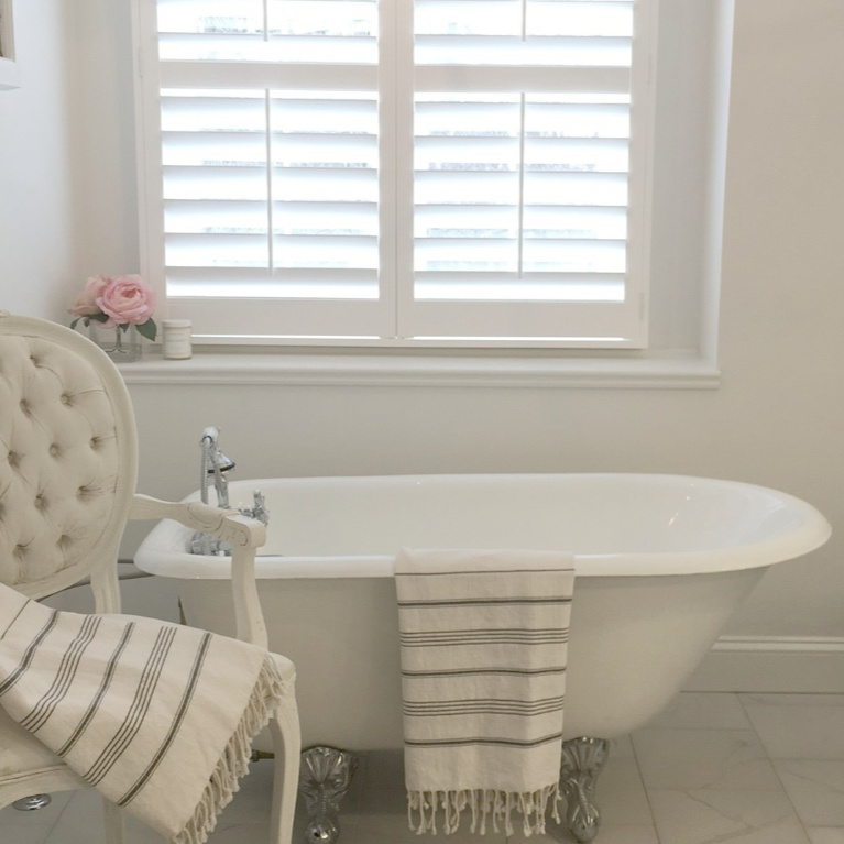 Clawfoot tub, French chair, and stripe Turkish towels in white French country bathroom by Hello Lovely Studio. #bathroomdesign #frenchcountry #hellolovelystudio #clawfoottub #plantationshutters #whitebathroom #frenchbathroom #vintagestyle