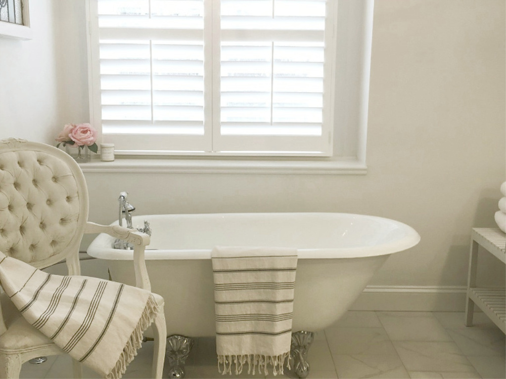 Clawfoot tub, French chair, and stripe Turkish towels in white French country bathroom by Hello Lovely Studio. #bathroomdesign #frenchcountry #hellolovelystudio #clawfoottub #plantationshutters #whitebathroom #frenchbathroom #vintagestyle