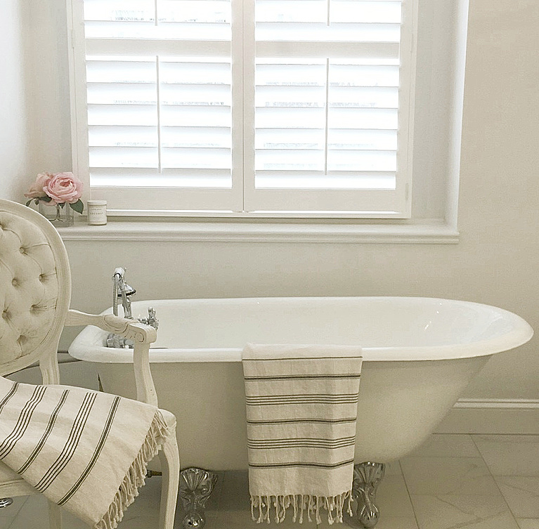 Clawfoot tub, French chair, and stripe Turkish towels in white French country bathroom by Hello Lovely Studio. #bathroomdesign #frenchcountry #hellolovelystudio #clawfoottub #plantationshutters #whitebathroom #frenchbathroom #vintagestyle