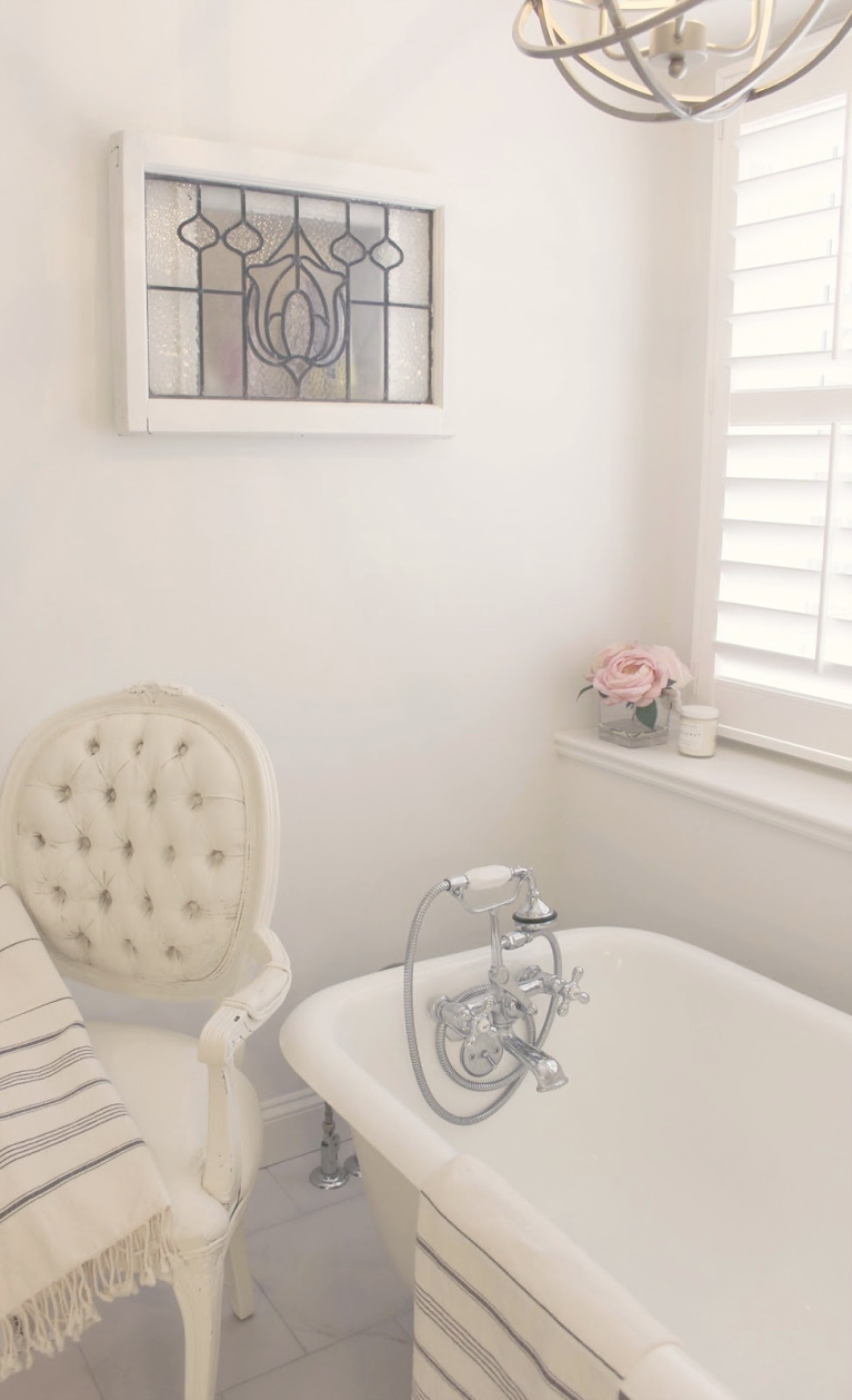 Clawfoot tub, French chair, and stripe Turkish towels in white French country bathroom by Hello Lovely Studio. #bathroomdesign #frenchcountry #hellolovelystudio #clawfoottub #plantationshutters #whitebathroom #frenchbathroom #vintagestyle