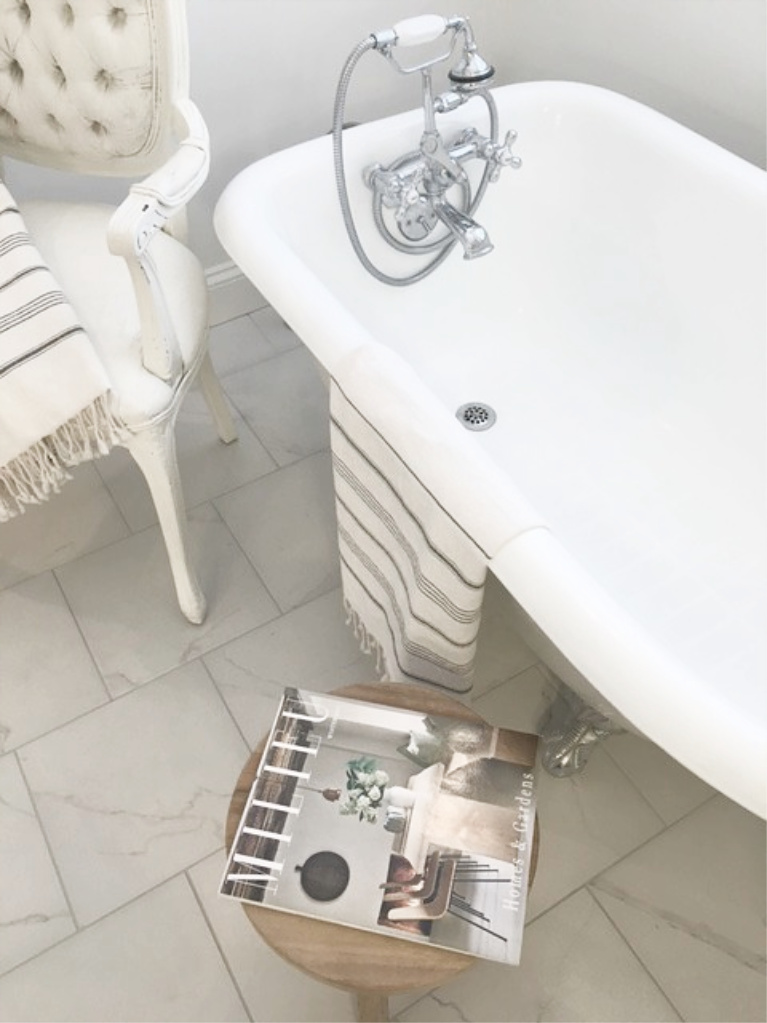 Clawfoot tub, French chair, and stripe Turkish towels in white French country bathroom by Hello Lovely Studio. #bathroomdesign #frenchcountry #hellolovelystudio #clawfoottub #plantationshutters #whitebathroom #frenchbathroom #vintagestyle