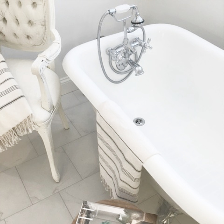 Clawfoot tub, French chair, and stripe Turkish towels in white French country bathroom by Hello Lovely Studio. #bathroomdesign #frenchcountry #hellolovelystudio #clawfoottub #plantationshutters #whitebathroom #frenchbathroom #vintagestyle