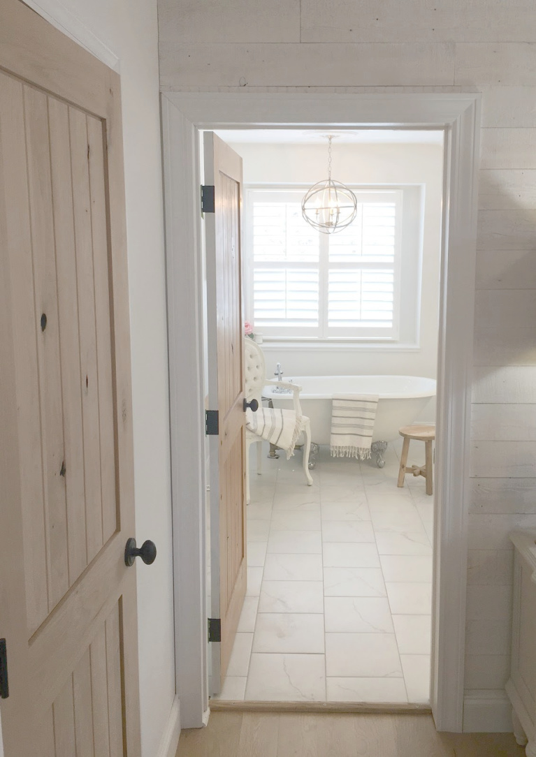 White French country master bathroom with rustic alder door, calacatta-look porcelain tile and clawfoot tub. #hellolovelystudio #bathroomdesign #bathroomdecor #frenchcountry #clawfoottub #alderdoor