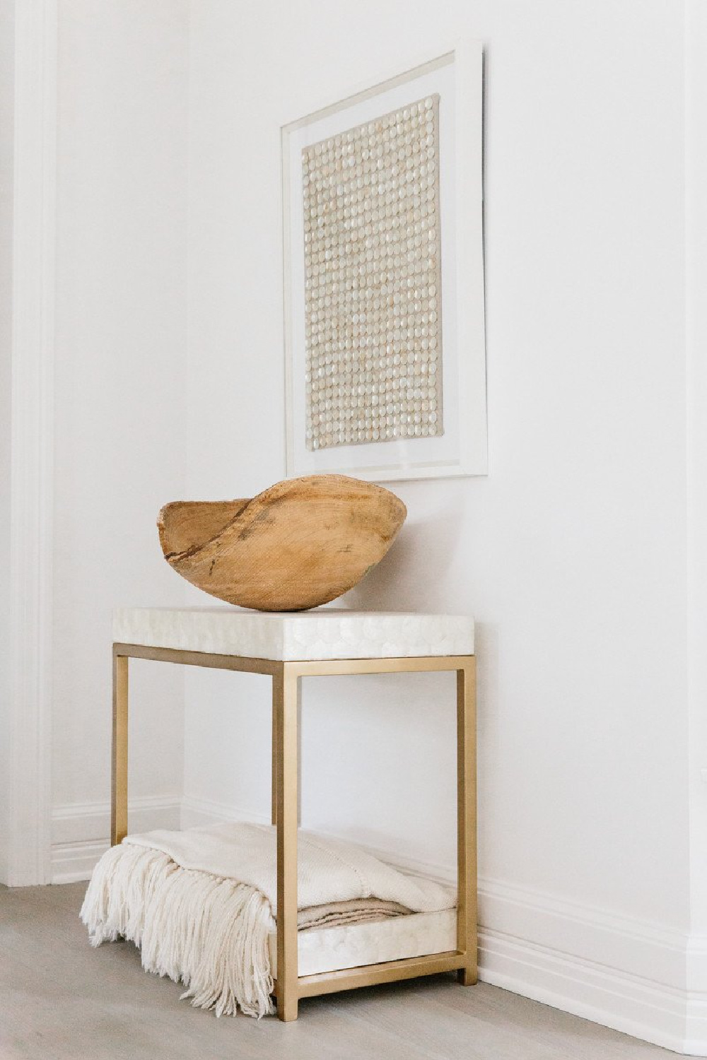 Mother of Pearl framed art over a marble and brass accent table topped with rustic wood dough bowl in Erin Fetherston's white on white living room in LA.