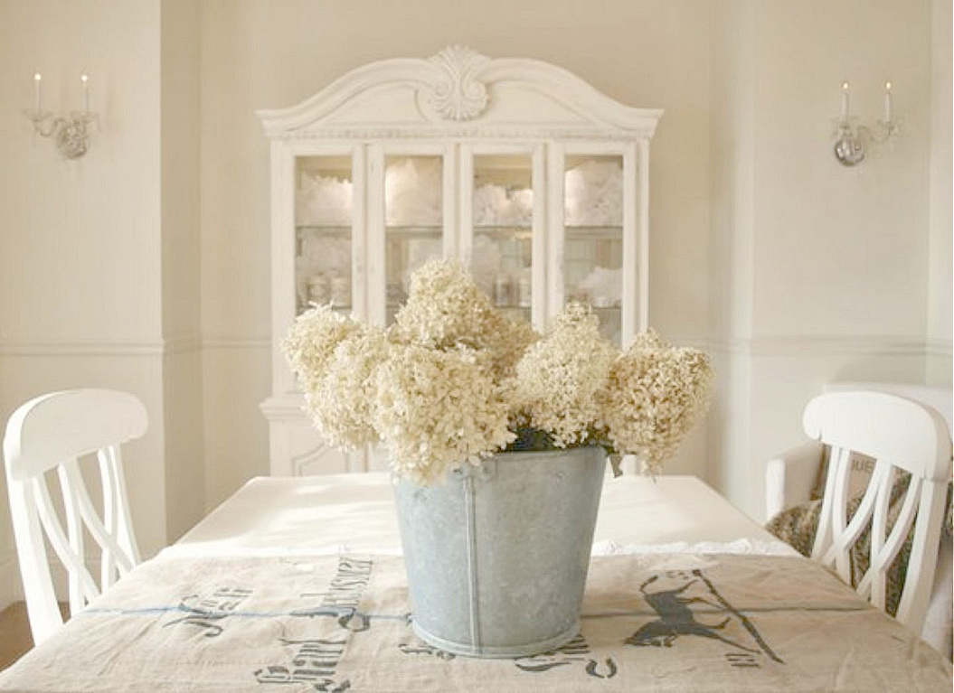 White French Nordic dining room with German antique grainsack and galvanized bucket of hydrangea on table - Hello Lovely Studio. #frenchnordic #diningroom #frenchcountry #whitefrench #grainsack #hydrangea #benjaminmoorewhitesand