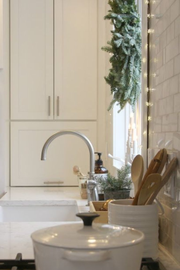 White Scandi inspired Christmas kitchen with fairy lights, fresh green wreath on window, and white decor - Hello Lovely Studio. #christmasdecor #whitechristmas #christmaskitchen #scandichristmas #serenechristmas #modernfarmhouse