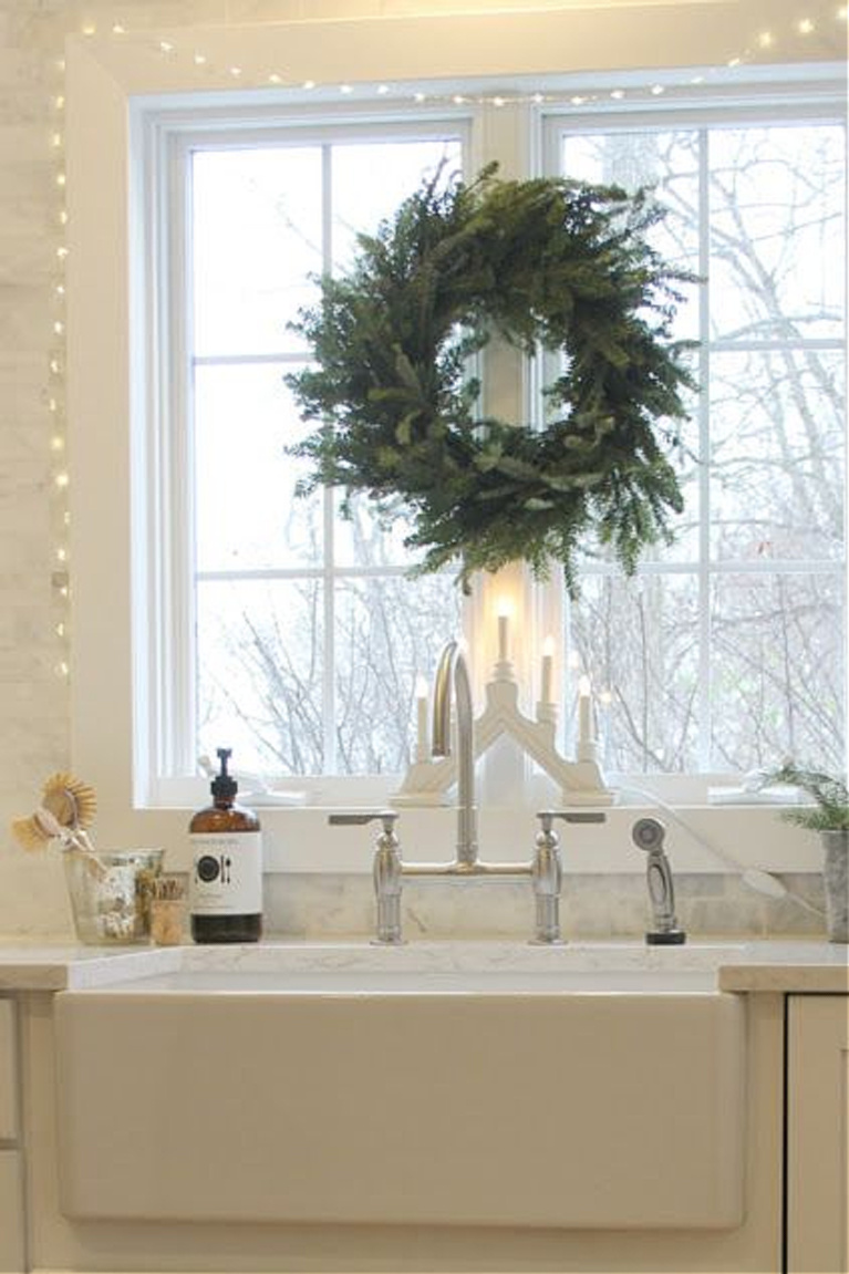 White Scandi Christmas decor in my farmhouse kitchen with Frasier fir wreath and Swedish candelabra at the farm sink in our white kitchen - Hello Lovely Studio. #christmasdecor #christmaswreath #hellolovelystudio #scandichristmas #christmaswreaths #candelabra #swedishchristmas