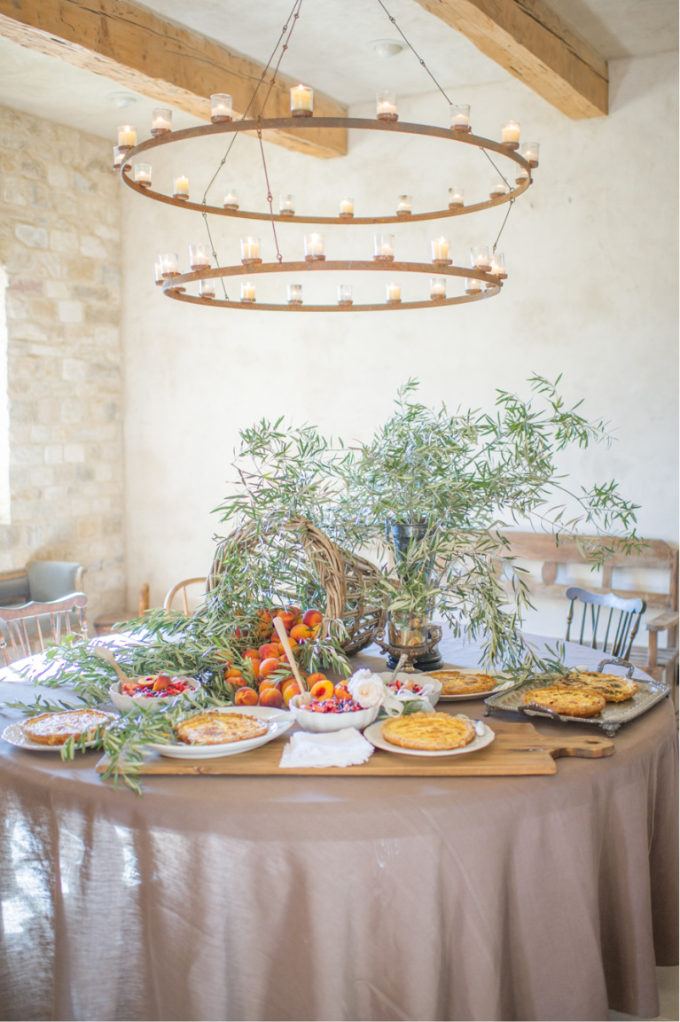 Rustic and elegant, this round French Country dining table features quiche, fresh fruit and olive branches with large ring candle pendants - Sunstone Winery - photo by Shannon Von Eschen. #frenchfarmhouse #tablescape #entertaining #frenchcountry