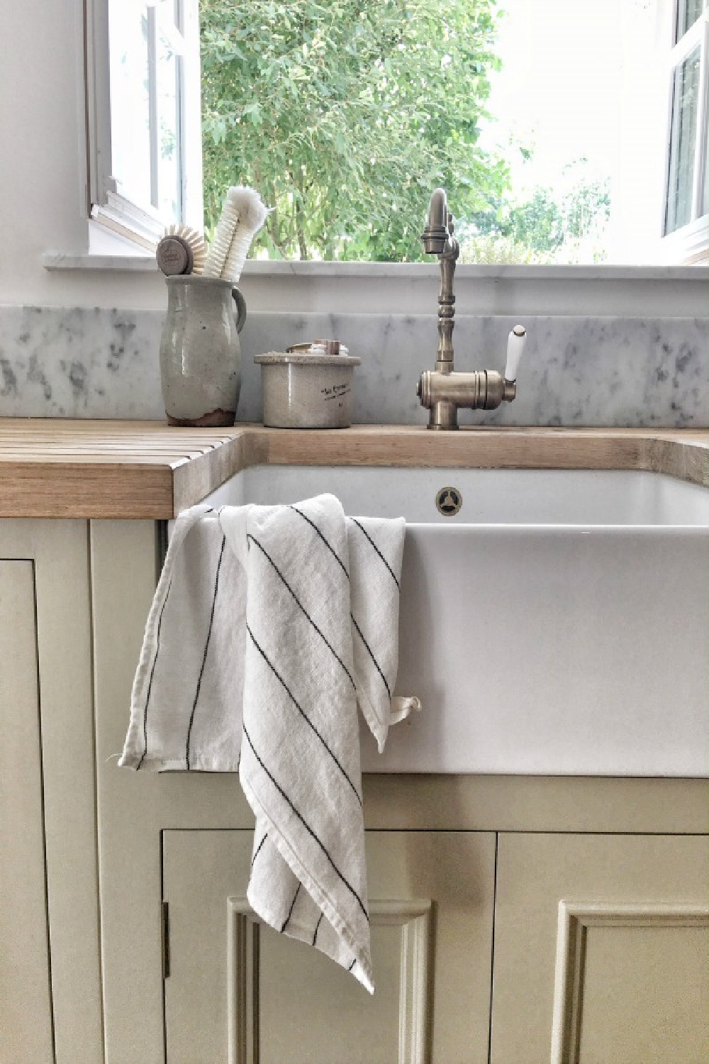 Detail of farm sink in a kitchen in France with putty colored cabinets, butcherblock countertop, marble backsplash, and brass faucet. #vivietmargot #frenchkitchens #farmsinks