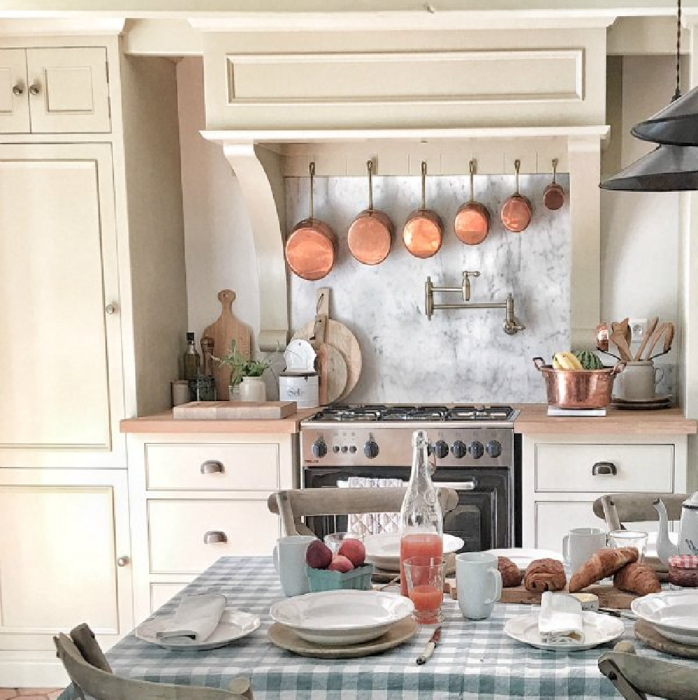 Charming check tablecloth, copper pans, and pale cabinets in French farmhouse kitchen by Vivi et Margot. 