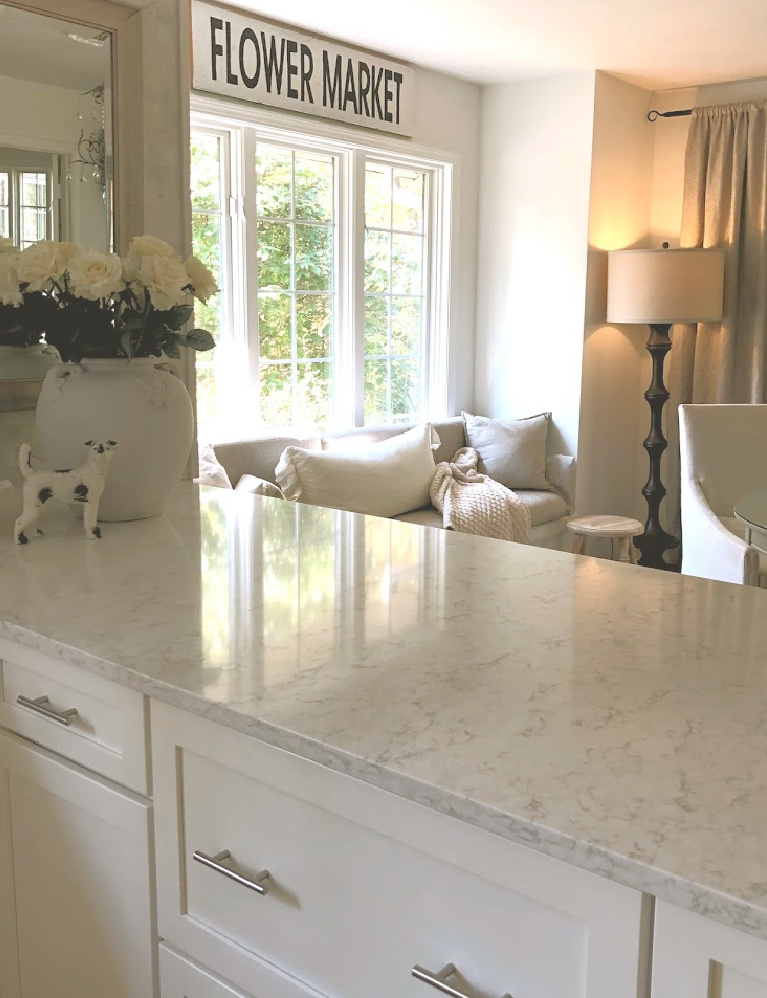 Serene white kitchen with LG Viatera Minuet quartz counters, Shaker cabinets, window seat, and white roses in French pot - Hello Lovely Studio. #hellolovelystudio #kitchendesign #lgviatera #minuet #whitequartz #windowseat #frenchcountry #europeancountry