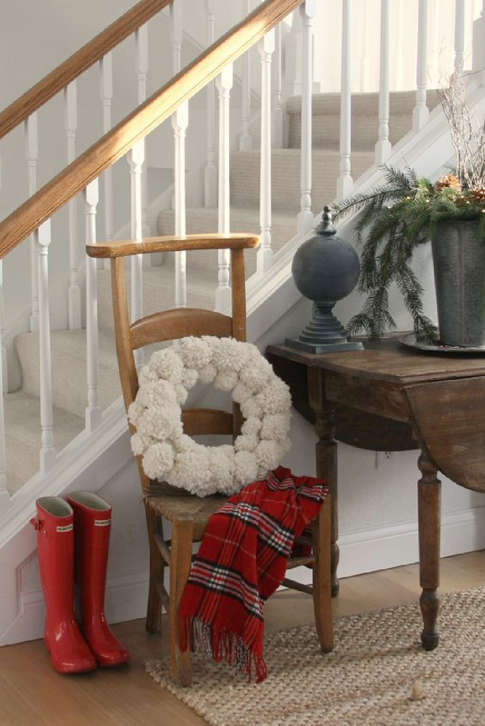 Christmas decor in a country house entry with red boots, red plaid scarf, wooly pom pom wreath, and fresh greenery. #hellolovelystudio #christmasdecor #redboots #pompomwreath