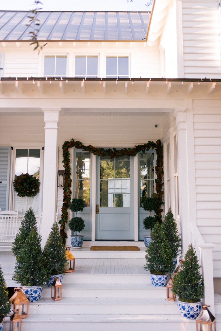 Charming, traditional and classic white and blue porch decorated for Christmas with magnolia garland, mini trees in porcelain planters and topiaries - Julia Berolzheimer.