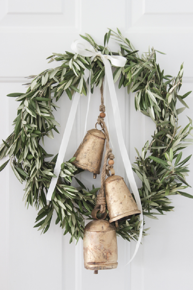 Gorgeous and minimal olive branch Christmas wreath with gold toned bells and white ribbon on a white door - Shades of Blue Interiors.